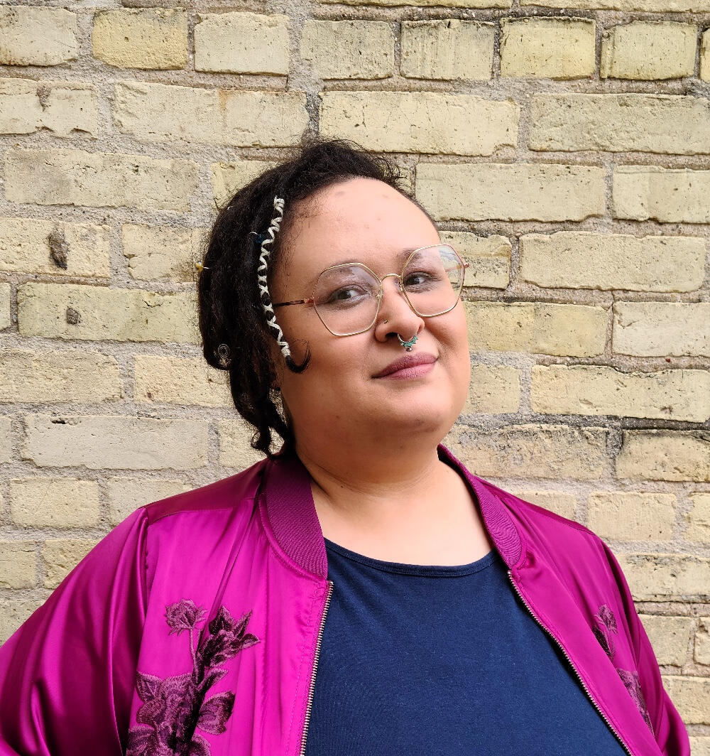 a light skinned black person in a fuchsia bomber jacket smiles while standing in front of a yellow brick wall.